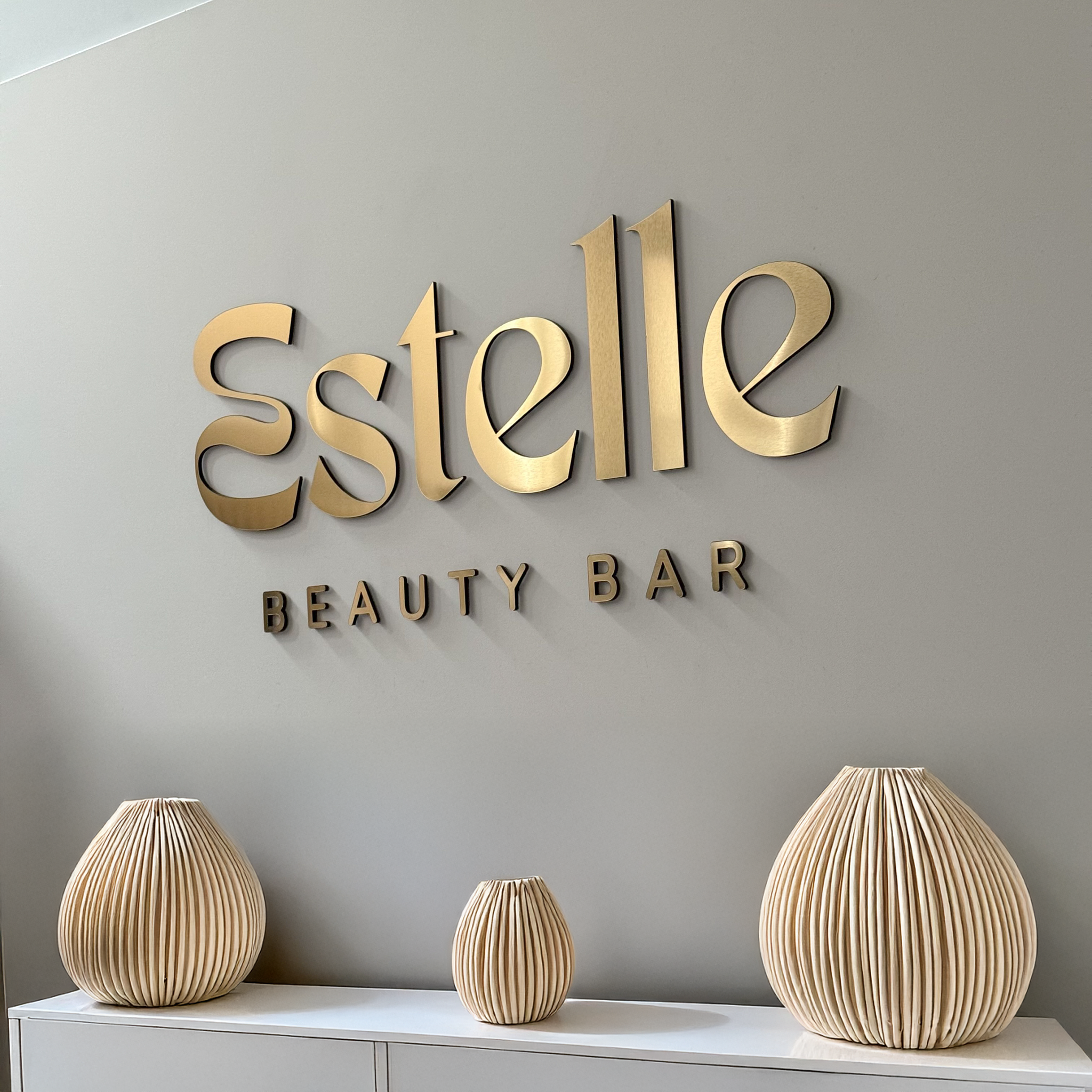 Gold 'Estelle Beauty Bar' sign on a gray wall with decorative vases below.